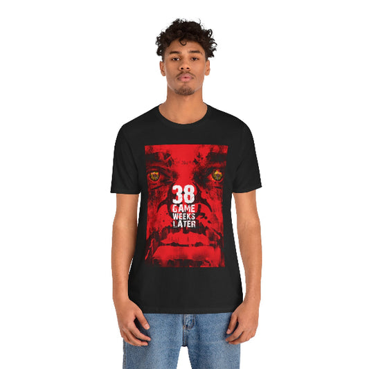 A man wearing a black short-sleeve t-shirt with a red graphic print on the front that reads '38 GAME WEEKS LATER' in white text, paired with blue jeans, against a plain white background.