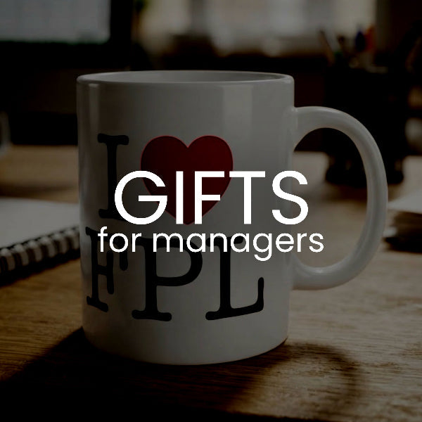 A white ceramic mug on a wooden desk with a dark overlay reading 'GIFTS for managers'. The mug features a black graphic of the letters 'I', 'F', 'P', 'L' surrounding a red heart.