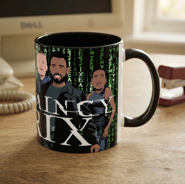 A ceramic mug with a black handle and interior, printed with a Matrix-style green code background, illustrated characters, and the text 'JANICE RIX'. The mug sits on a wooden desk with office items blurred in the background.