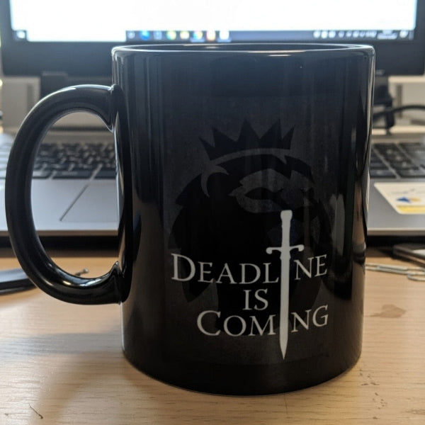 Black mug with 'Deadline is Coming' design on a desk with a computer in the background