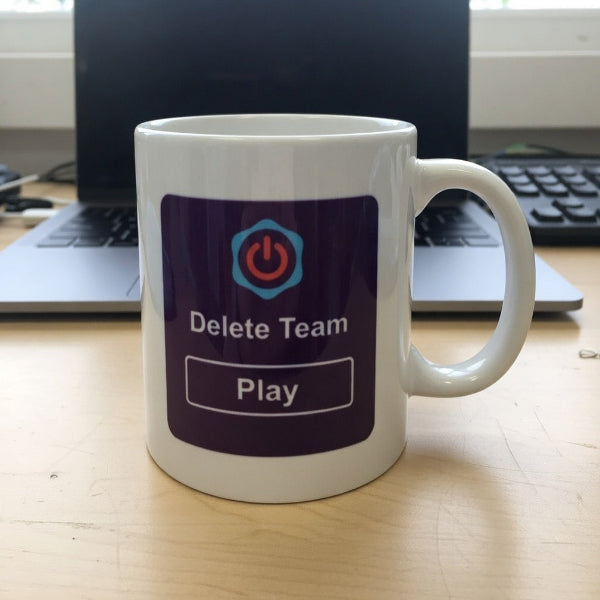 White mug with 'Delete Team Play' design on a desk with a laptop in the background
