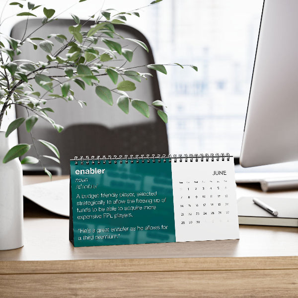 A spiral-bound desk calendar showing June with a teal and white page titled 'enabler' sits on a wooden desk in an office setting, with a plant, chair, and computer monitor blurred in the background.