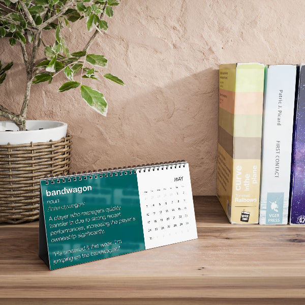 A spiral-bound desk calendar standing on a wooden surface, with a teal and white page showing the word 'bandwagon' and a May calendar grid. The calendar is styled in a home office setting next to a potted plant and books against a textured beige wall.
