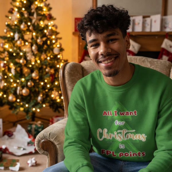 A smiling man wearing a bright green crewneck sweatshirt with the text 'All I want for Christmas is FPL points' in red and white lettering, seated in a cozy living room with a decorated Christmas tree in the background.