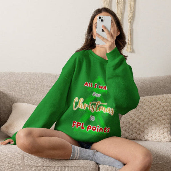 A woman sits on a beige couch taking a mirror selfie while wearing a bright green sweatshirt with a humorous graphic that reads 'All I want for Christmas is FPL points' in red and yellow text.