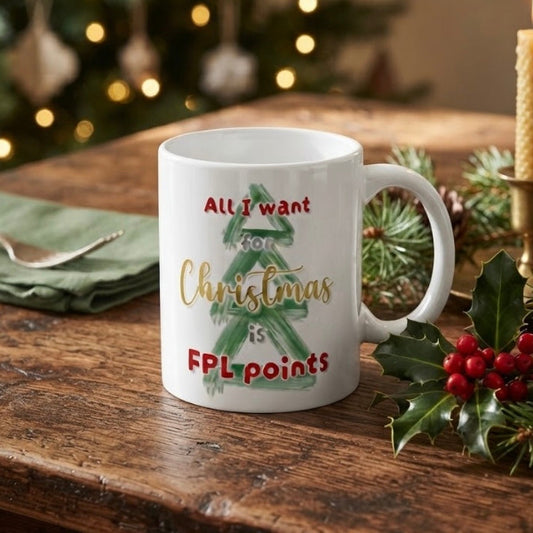 A white ceramic Christmas mug with a green tree graphic and the text 'All I want for Christmas is FPL points', styled on a rustic wooden table with holiday greenery and warm blurred lights in the background.