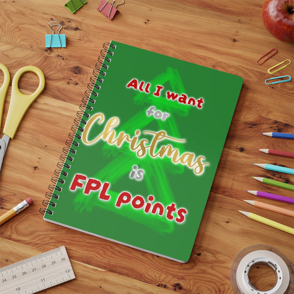 A green spiral-bound notebook with a Christmas-themed quote, displayed on a wooden desk surrounded by school and office supplies.