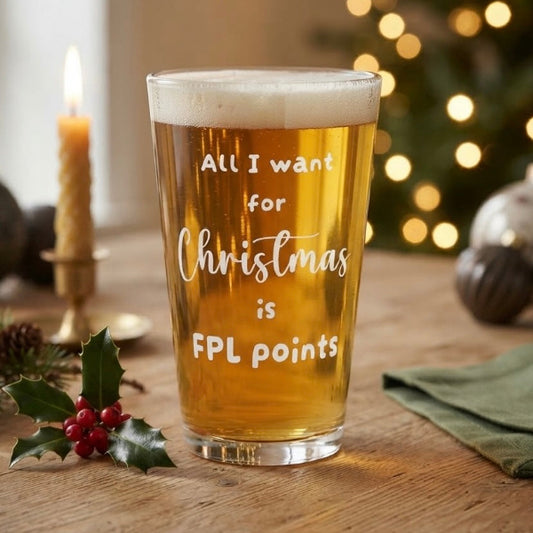 A frosted pint glass filled with golden beer, standing on a wooden table. The glass has white text that reads 'All I want for Christmas is FPL points'.