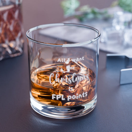 A clear glass rocks tumbler containing amber whiskey, with white frosted text reading 'All I want for Christmas is FPL points'. The glass is photographed on a dark tabletop with a softly blurred indoor background.
