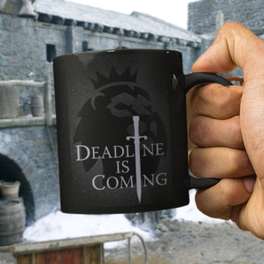 Black mug with 'Deadline is Coming' text held by a hand in front of a stone building.
