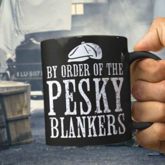 Black mug with 'By Order of the Pesky Blankers' text held by a hand, with a blurred background.