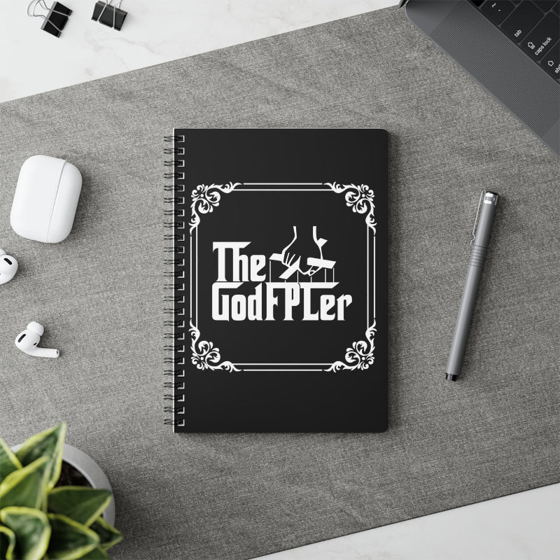 Black notebook with 'The GodFPLer' design on a desk with a pen and earbuds.