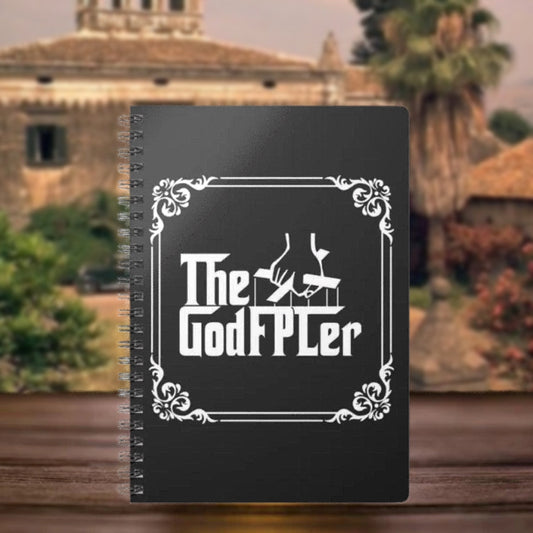 Black notebook with 'The GodFPLer' design on a blurred background of a building and trees