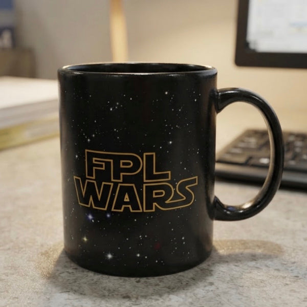 A black ceramic coffee mug with a starry space pattern and the text 'FPL WARS' in a gold Star Wars-style font, sitting on a countertop with a blurred office background.