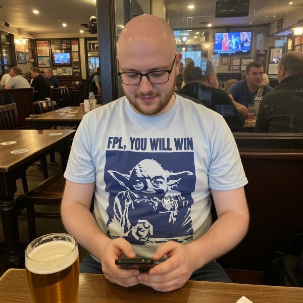 Man sitting at a bar using a phone with a beer in front of him, wearing a t-shirt with a graphic design.
