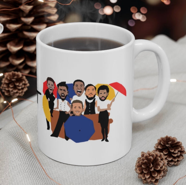 A white ceramic coffee mug filled with black coffee, decorated with a colorful cartoon illustration of five men. The mug is set on a light beige textured fabric surrounded by pinecones and warm string lights.