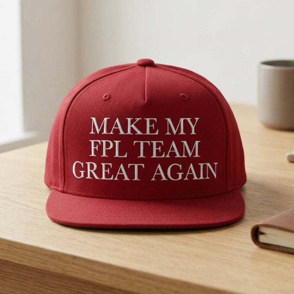 A red flat-brim baseball cap with white embroidered text reading 'MAKE MY FPL TEAM GREAT AGAIN' sits on a light wood table in a softly lit indoor setting.