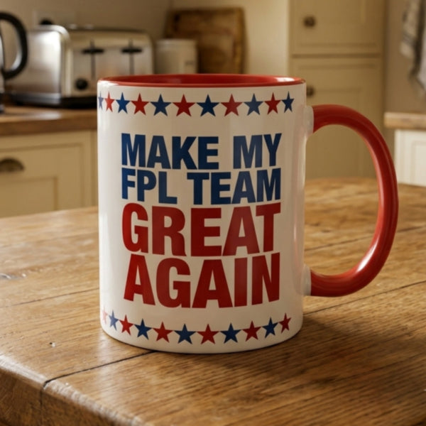 A white ceramic mug with a red handle and red interior, printed with the slogan 'MAKE MY FPL TEAM GREAT AGAIN' in blue and red text, surrounded by red and blue stars, sitting on a wooden table in a kitchen setting.