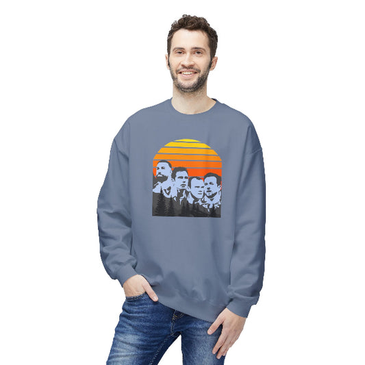 A man wearing a slate blue crewneck sweatshirt with a retro graphic of four men's faces in front of a yellow and orange sunset, paired with blue jeans, on a solid white background.