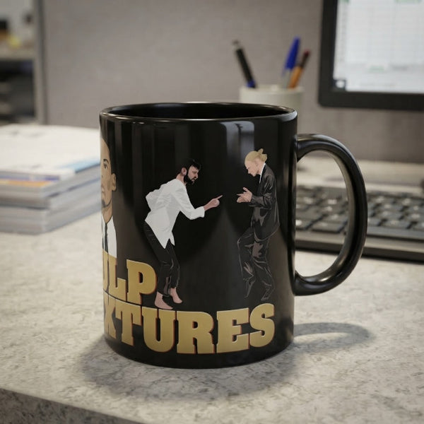 A black ceramic mug with a glossy finish, featuring a cartoon caricature of a man with a beard and blonde hair, and the text 'HELP TITLES' in gold. The mug is sitting on a gray speckled countertop in an office environment with a keyboard and computer monitor blurred in the background.