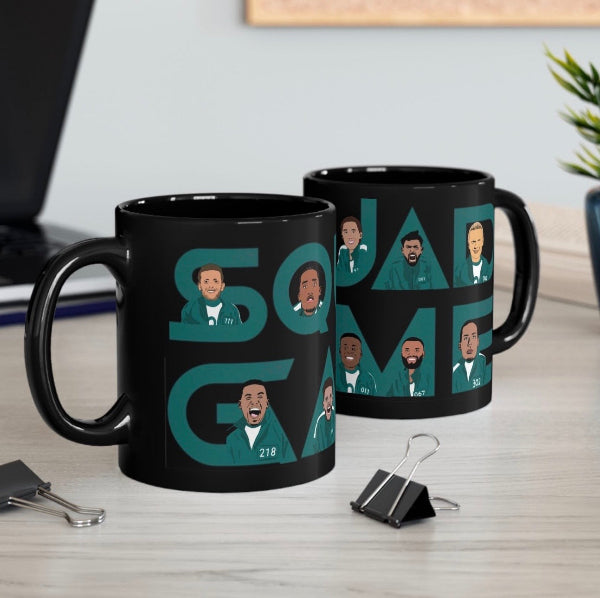 Two black ceramic mugs printed with a stylized 'Squid Game' design, displayed on a light wood desk with binder clips and a laptop in the background.