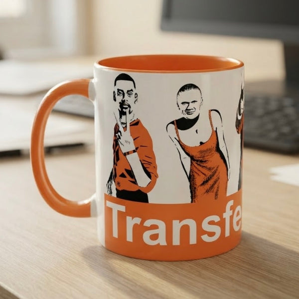 An orange and white ceramic mug featuring a graphic print of the band Transfer with their name printed in white on an orange band at the bottom, sitting on a wooden desk with a blurred office background.