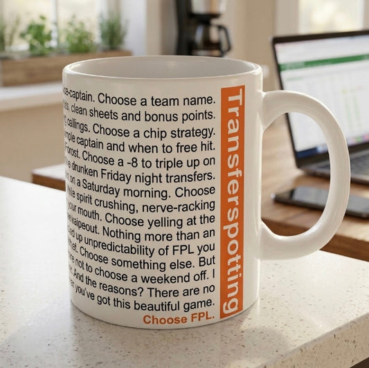 A white ceramic mug with black text and an orange vertical 'Transferspottin'' label, sitting on a kitchen countertop with a blurred laptop in the background.