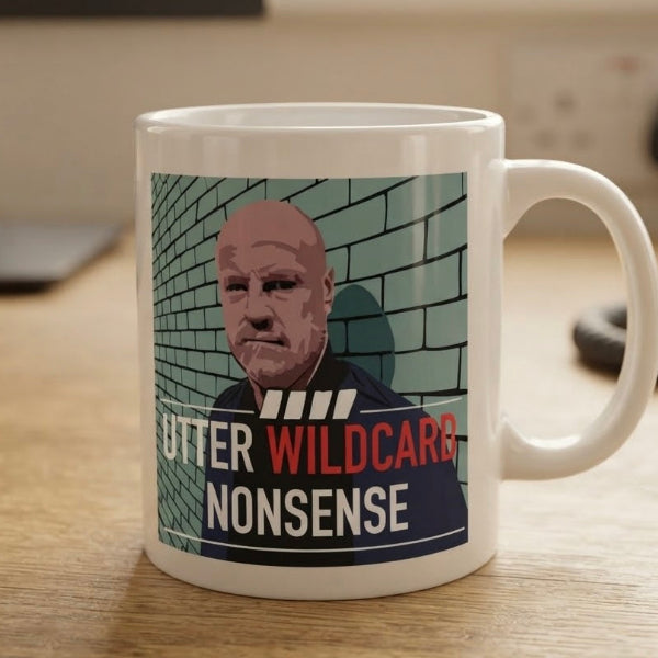 A white ceramic mug with a printed graphic of a bald man in front of a brick wall, with the text 'UTTER WILDCARD NONSENSE' in red and white. The mug is sitting on a wooden tabletop with a blurred indoor background.